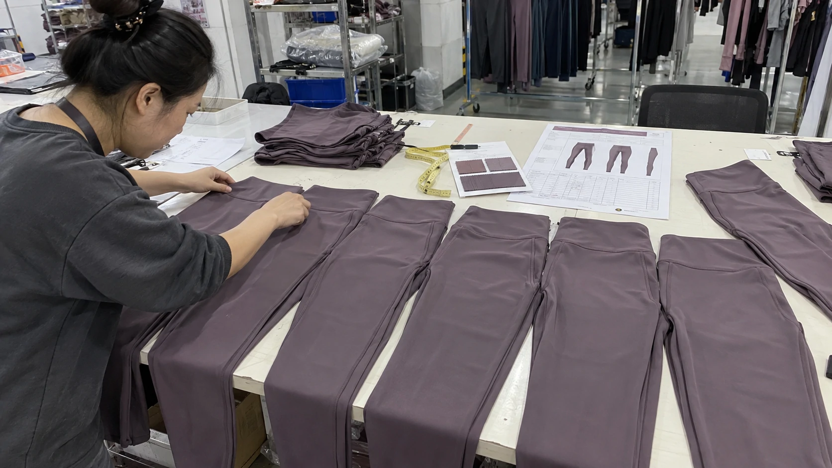 Worker inspecting multiple mauve activewear leggings against approved samples, fabric swatches and a tech pack during bulk production quality control